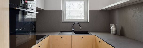 Panorama of elegant kitchen with birch plywood on furniture fronts, dark veneer countertops, oven, black sink with tap and one small window - 写真・画像