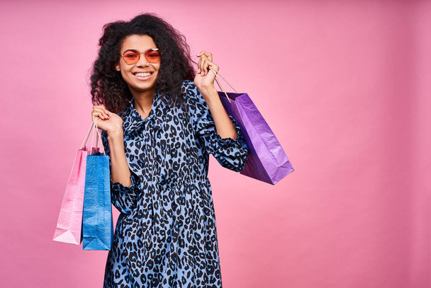Dreamy attractive young dark-skinned woman with curly hair, wearing red sunglasses, and a sincere happy smile, holding a bunch of shopping bags. Copy space. - Photo, image