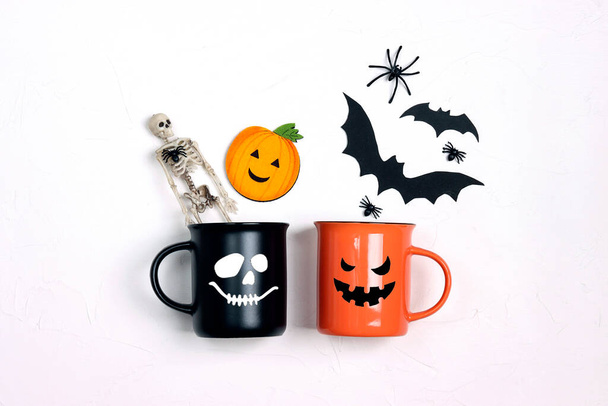 Black and orange mugs with funny faces and Halloween decorations on white table. Festive Halloween decorations and drinks. - Foto, imagen