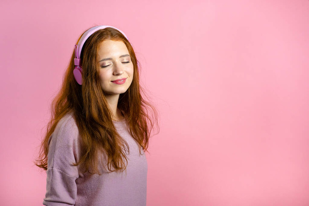Aantrekkelijke vrouw met rood haar met hoofdtelefoon op roze studioachtergrond. Schattig meisjesportret. Muziek, radio, geluk, vrijheid, jeugdconcept. - Foto, afbeelding