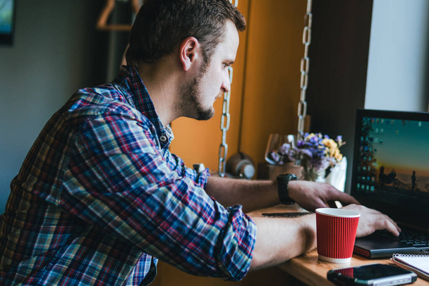 Ekose gömlekli adam elinde sıcak kırmızı bir fincan çay tutuyor. İş yeri, dizüstü bilgisayarı ve kalemi olan defter. Kahve dükkanı, mesaj için yer, bilgisayarın başında sabah işi, ders çalışma. - Fotoğraf, Görsel