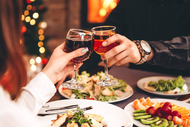 Close up of hands holding wine glasses and clinking against the backdrop of bright Christmas tree and burning fireplace, couple celebrating at Christmas table - Φωτογραφία, εικόνα