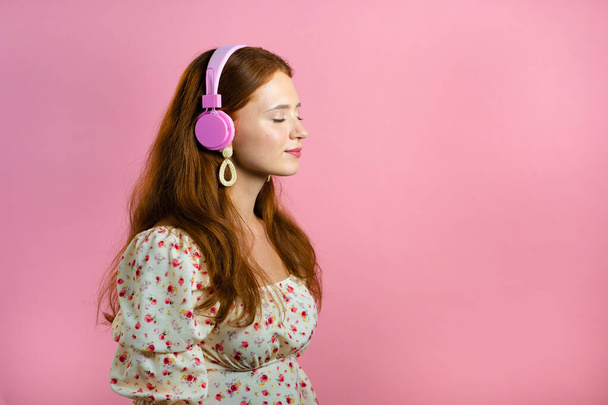 Attractive woman with red hair with headphones on pink studio background. Cute girl portrait. Music, radio, happiness, freedom, youth concept. - Photo, image