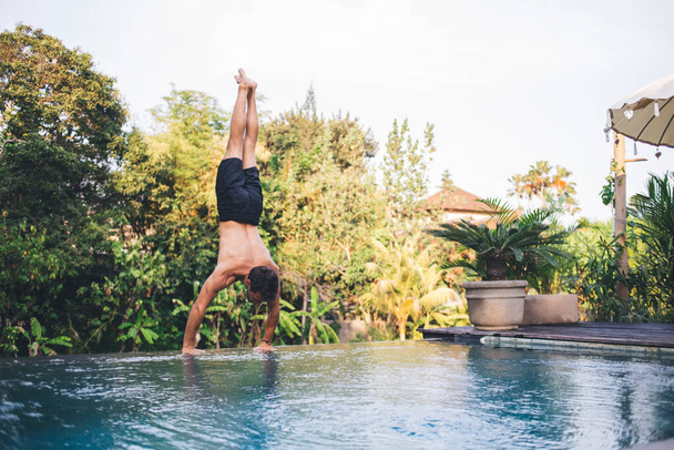 Back view of unrecognizable fit young male athlete with naked torso performing handstand asana near outdoor pool against green trees while doing yoga in Bali - Foto, immagini