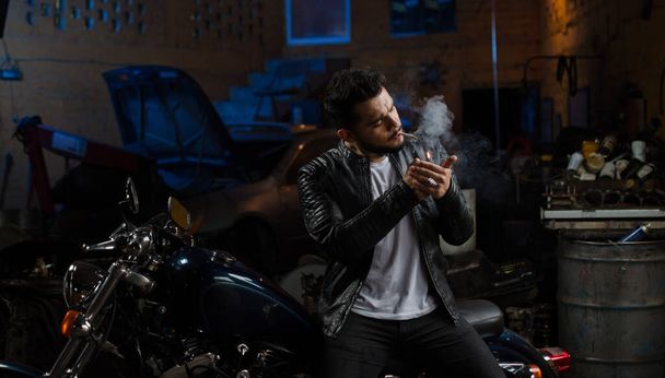 A male sitting on a motorcycle in his garage lighting a cigarette - Photo, image