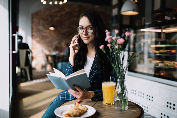 Glad young lady in glasses with book sitting at table with food and vase of flowers while talking on phone and looking at camera - Fotó, kép