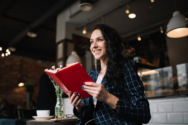 Od dołu zadowolona kobieta w stylowym stroju uśmiecha się i odwraca wzrok czytając ciekawą książkę podczas przerwy na kawę w kawiarni - Zdjęcie, obraz