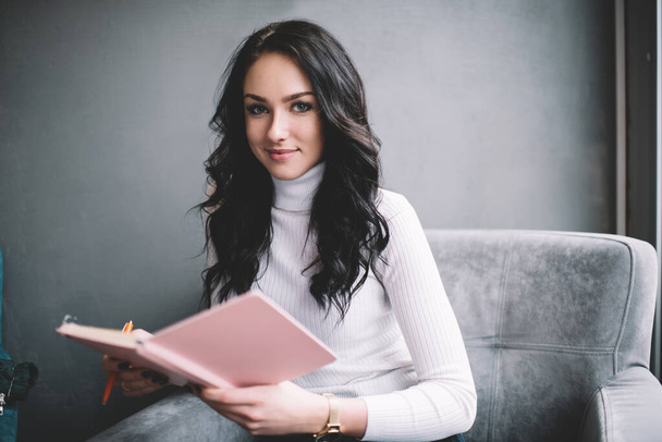 Positive female entrepreneur wearing casual clothes sitting in comfortable armchair with pink notebook and looking at camera while thinking about business - Φωτογραφία, εικόνα