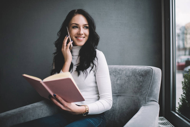 Young female entrepreneur wearing casual clothes sitting in comfortable armchair and speaking on smartphone while checking information in notepad and looking away - Foto, Imagem