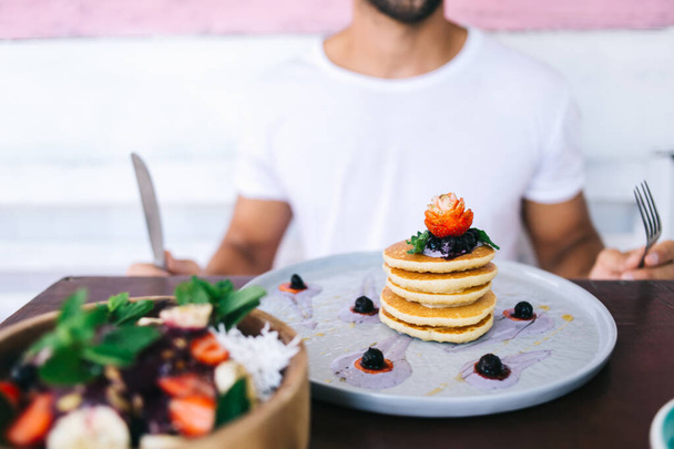 Unrecognizable crop man sitting at table with fork and knife and preparing eating pancakes decorated with strawberry and berries - Foto, imagen