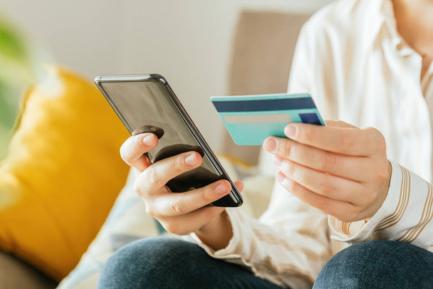 Stock photo of an unrecognizable young woman shopping online with the phone and a credit card from the sofa at home. E-commerce concept - Photo, Image