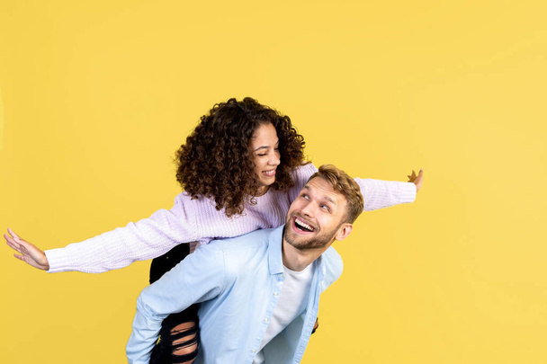 Happy adult man carry smiling african american woman while she making airplane gestures. Two friends having fun, standing isolated on yellow copy space background - Photo, Image