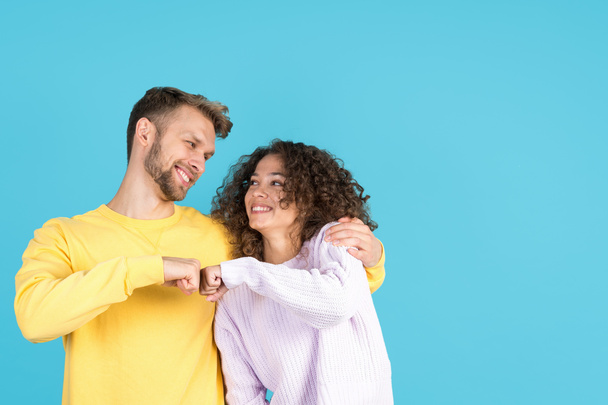 Happy mixed race friends bump with fists, making greeting gesture, hugging and smiling wide while they standing isolated on blue copy space background - Photo, Image