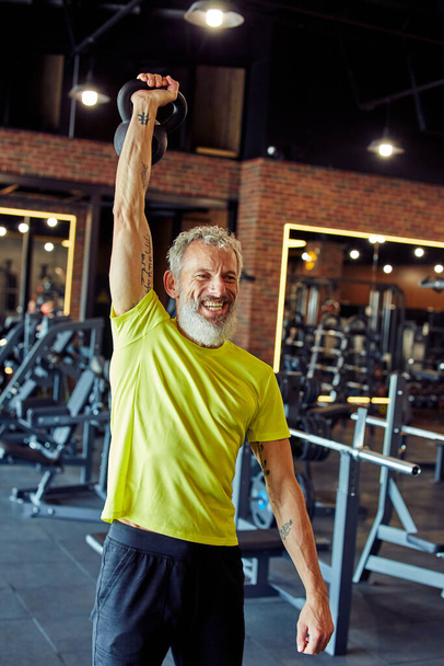 Вертикальный снимок улыбающегося взрослого кавказца в спортивной форме с гантелями в спортзале - Фото, изображение
