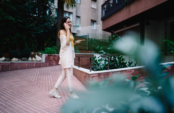 Side view of full body positive young lady in casual summer wear and eyeglasses speaking on smartphone near entrance of building - Фото, зображення