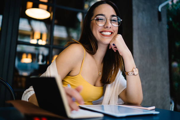 Smiling young female entrepreneur in eyeglasses and casual clothes working with notebook and tablet while sitting at table in modern outdoor cafe and looking at camera - Foto, Imagen