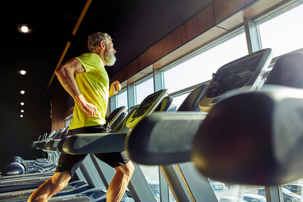 Seitenansicht eines athletischen Mannes mittleren Alters in Sportbekleidung, der in einem Fitnessstudio auf einem Laufband läuft und vor dem Training Cardio macht - Foto, Bild
