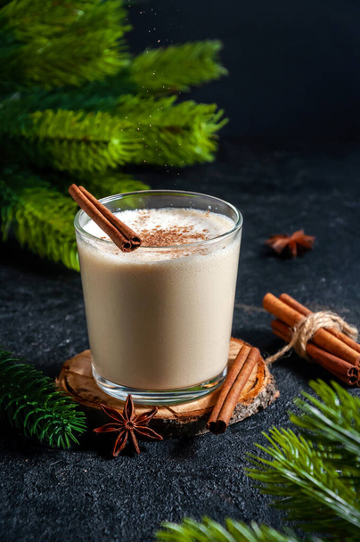 Christmas Hot Drink. Eggnog with Sifting Cinnamon in Glass with Branches Fir Tree on Dark Background. - Photo, Image