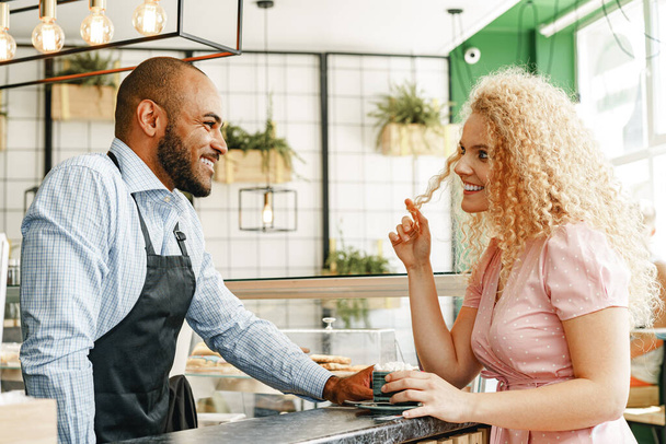 Smiling blonde woman talking to a waiter of a coffee shop at the counter - Foto, Bild