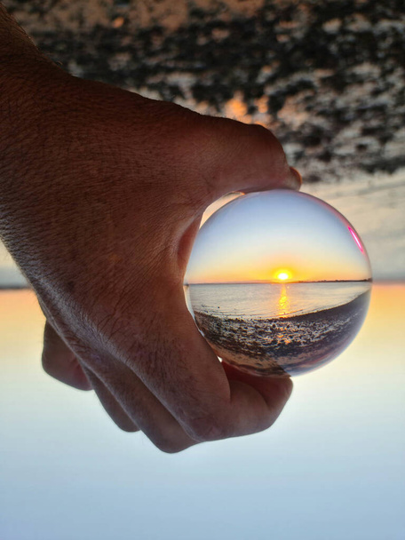 Gün batımında sahili yansıtan cam küreyi tutan bir adamın dikey görüntüsü. - Fotoğraf, Görsel