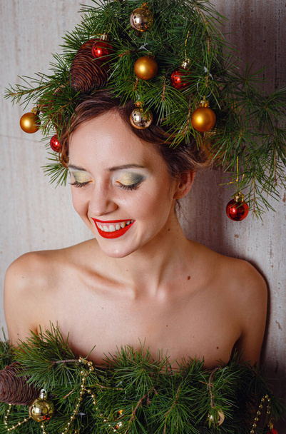 Piękna młoda dama ze świątecznym wieńcem sosnowym na głowie i modną sukienką na choinkę, ozdobioną świątecznymi kulkami, jagodami i płatkami śniegu. Koncepcja nowego roku - Zdjęcie, obraz