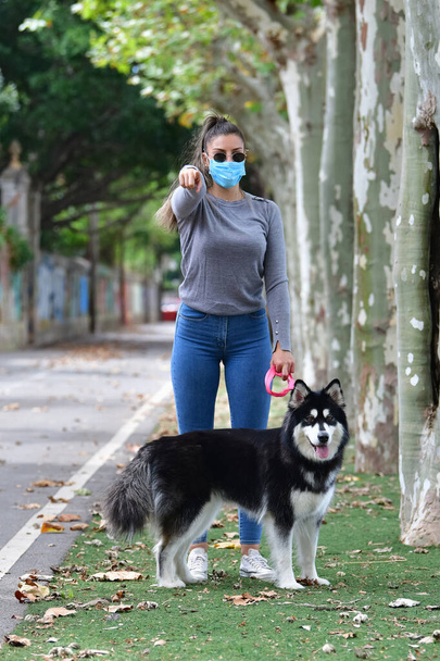 Woman with glasses and surgical mask holding a dog on a leash pointing towards the camera on an out of focus background. New normal and pets concept. - Fotografie, Obrázek