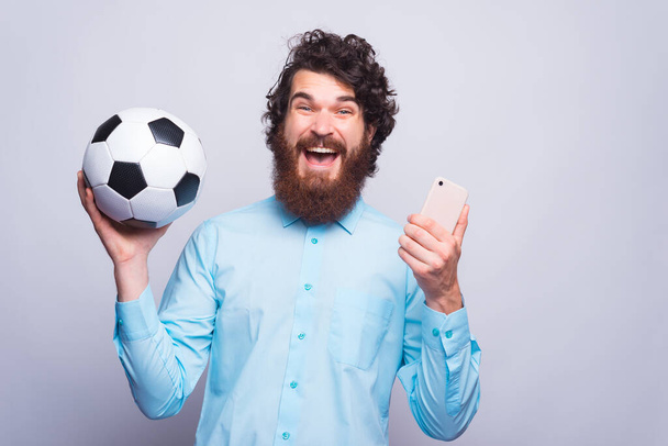 Νεαρός μουσάτος χίπστερ ενθουσιασμένος κρατά ένα κινητό τηλέφωνο και μια μπάλα ποδοσφαίρου κοντά σε ένα λευκό τοίχο - Φωτογραφία, εικόνα