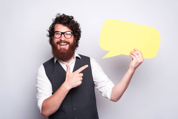 Bearded young hipster with glasses is smiling at the camera and holding a speech bubble and pointing at it near a whtie wall - Φωτογραφία, εικόνα