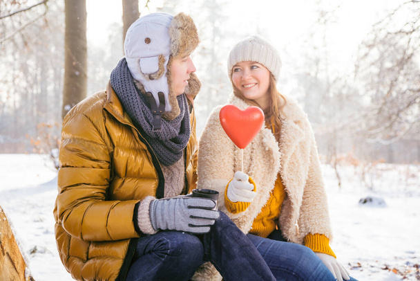 mladý pár drží červené srdce a usmívají se navzájem, zatímco sedí spolu v zimním parku   - Fotografie, Obrázek