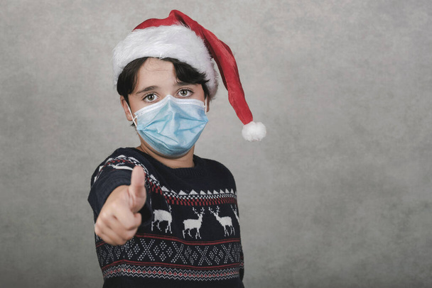 Veselé Vánoce, dítě s lékařskou maskou na sobě Santa Claus klobouk ukazující palec nahoru přes šedé pozadí - Fotografie, Obrázek