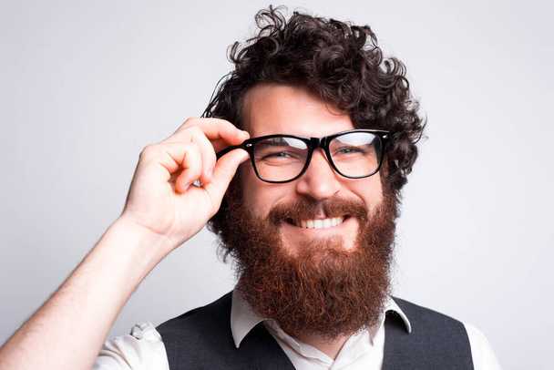 Фото молодого бородатого хипстера в очках, улыбающегося в камеру возле белой стены - Фото, изображение
