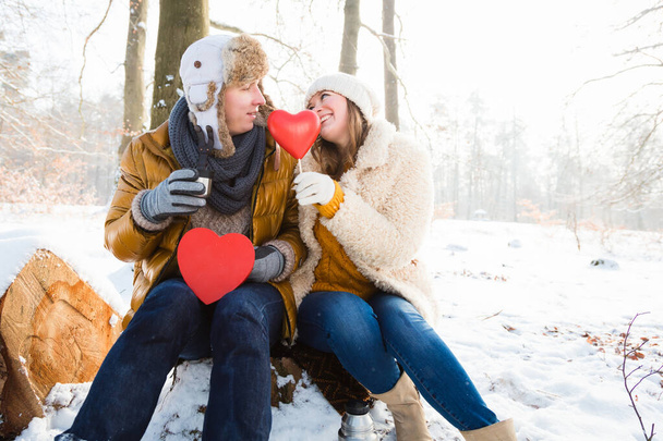 mladý pár drží červená srdce a usmívají se navzájem, zatímco sedí spolu v zimním parku   - Fotografie, Obrázek