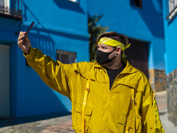 A young Caucasian male with a yellow coat and a medical mask taking a selfie with a phone in Spain- the new normal due to Covid-19 - Фото, зображення