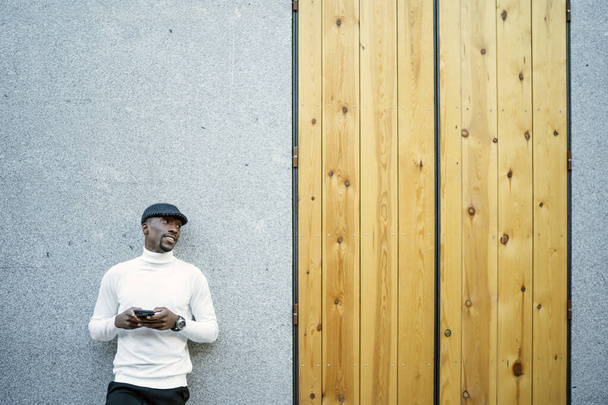 A closeup shot of a black man wearing a hat and a turtleneck holding his phone - Zdjęcie, obraz