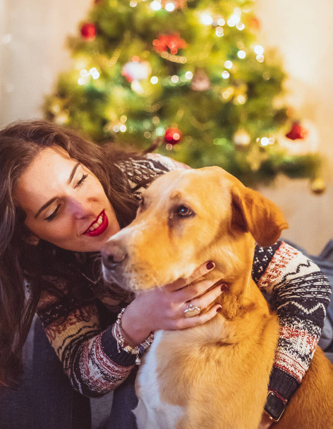 A vertical shot of a Caucasian woman hugging her dog with a Christmas tree on the background - Fotoğraf, Görsel