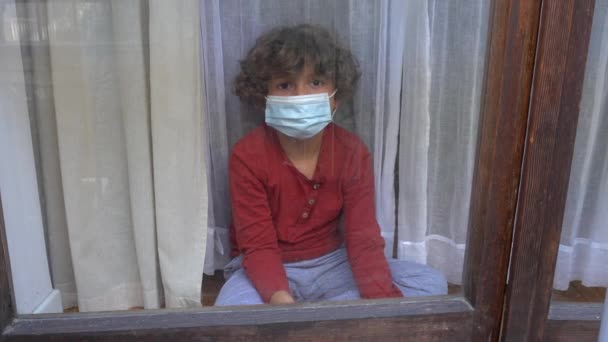 blanke blanke blanke jongen 6 jaar oud in quarantaine huis met masker kijkt uit het raam van het huis tijdens Coronavirus lockdown in Europa, Amerika en Azië - Video
