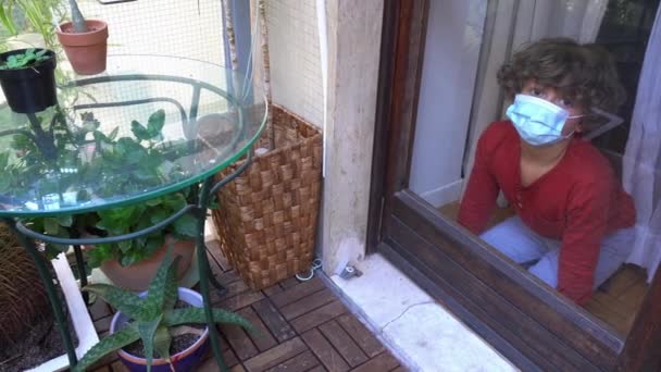 blanco caucásico niño 6 años en cuarentena casa con máscara mira por la ventana de la casa durante el bloqueo del Coronavirus en Europa, América y Asia - Metraje, vídeo