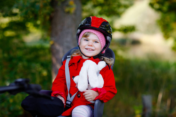 Маленька дівчинка з шоломом безпеки на голові сидить на велосипедному сидінні своєї матері або батька велосипеда. Безпечна концепція захисту дітей. Сімейна поїздка в осінній день. Малюк тримає поштовх м'якої іграшки
 - Фото, зображення