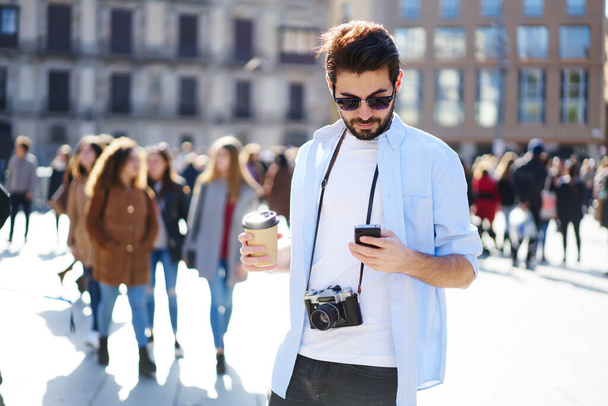 Homem barbudo sério em roupas casuais e óculos de sol com câmera de foto em pé na rua com xícara de café surf smartphone - Foto, Imagem