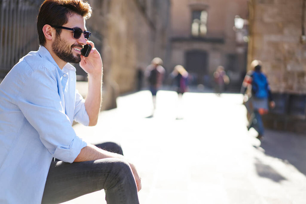 Side view of cheerful young man with beard in casual clothing and sunglasses having conversation on phone and looking away with bright smile - Fotoğraf, Görsel