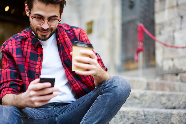 Crop focused young bearded male in casual clothes and eyeglasses looking at screen and messaging on cellphone while sitting on stone stairs and holding coffee to go - Zdjęcie, obraz