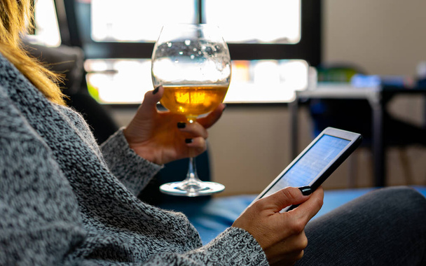 A woman with painted nails, holds with her right hand in the foreground an e-book and with her left hand a glass of beer, relaxed and calmly sitting on an armchair in her house, while light comes in through the windows in the back of her house - Photo, Image