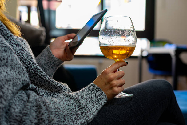 Eine Frau, die in der einen Hand ein Glas Bier hält und in der anderen ein Smatphone, ein E-Book oder ein Tablet, all dies in einem Sessel, mit den Fenstern ihres Hauses im Hintergrund. - Foto, Bild