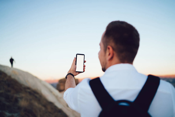 Back view of anonymous male in white clothes with black backpack using smartphone for taking selfie standing next to rock - Fotoğraf, Görsel
