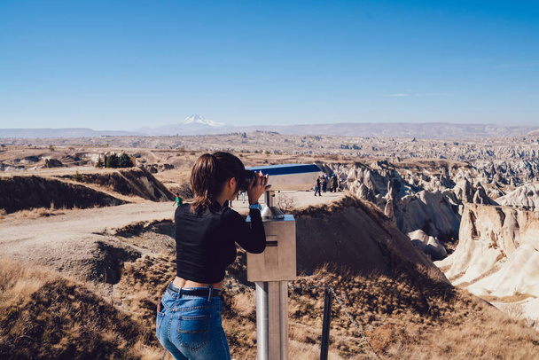 Back view of anonymous woman looking at rocky terrain through binoculars while standing on observation deck on sunny day in countryside - Foto, afbeelding