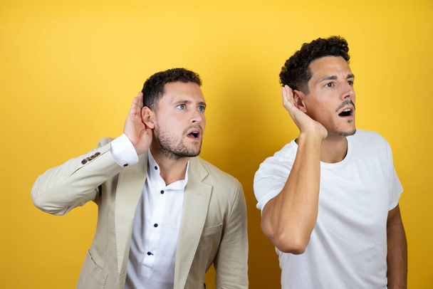 Junge schwule Paar von zwei Männern in lässiger Kleidung über isolierten gelben Hintergrund lächelnd mit der Hand über dem Ohr hören ein Gehör zu Gerüchten oder Klatsch - Foto, Bild