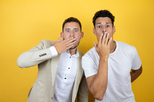Νεαρό γκέι ζευγάρι δύο ανδρών που φορούσαν καθημερινά ρούχα πάνω από απομονωμένο κίτρινο φόντο ξάφνιασε καλύπτοντας το στόμα της με το χέρι της. - Φωτογραφία, εικόνα