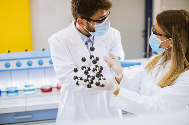 Νεαροί χημικοί με γυαλιά ασφαλείας κρατούν μοριακό μοντέλο στο εργαστήριο - Φωτογραφία, εικόνα