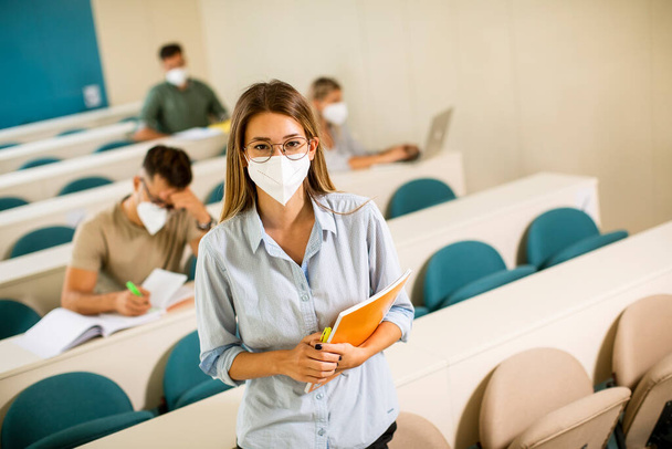 Young female student wearing face protective medical mask for virus protection standing at lecture hall - 写真・画像