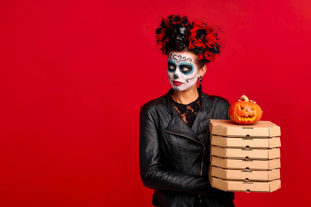Girl with sugar skull makeup, with a wreath of flowers on her head and skull, wearth lace gloves and leather jacket, holds pile of cardboard boxes with pizza. Delivery service and Halloween concept - Photo, Image